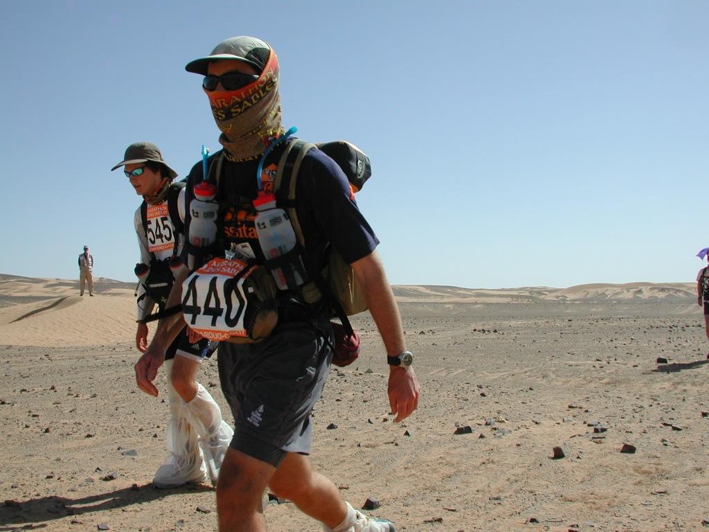 A side shot landscape photo of two people walking in the desert. They are participants of the Marathon Des Sables. A man in the centre is wearing a Original Buff® as face mask. A woman in the back is wearing a Original Buff® as neck cooler. It looks as if it is the end of the race and everybody is tired. The woman in the back is looking weak. The man in font still seems to have energy left. The photo was taken in 2003. Today the correct product for this race is the High UV Buff®. Source: buff.eu © Distributed for the promotion of the Original Buff® in outdoor / adventure