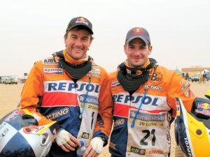 Frontal shot landscape photo of 2 motorcycle rallye bikers. It shows Marc Coma and Jordi Viladoms before the 6th etape of the 2007 Dakar Rally. They are both smiling into the camera. Interesting is the use of a High UV Buff® together with a neck brace. Look closer and you see the Buff® wrapping around the brace collar. That prevents rashes and keeps the sand out. It looks dusty and sandy in the background. Source: buff.eu Copyright: Distributed for the promotion of the High UV Buff® in motorcycle rallies