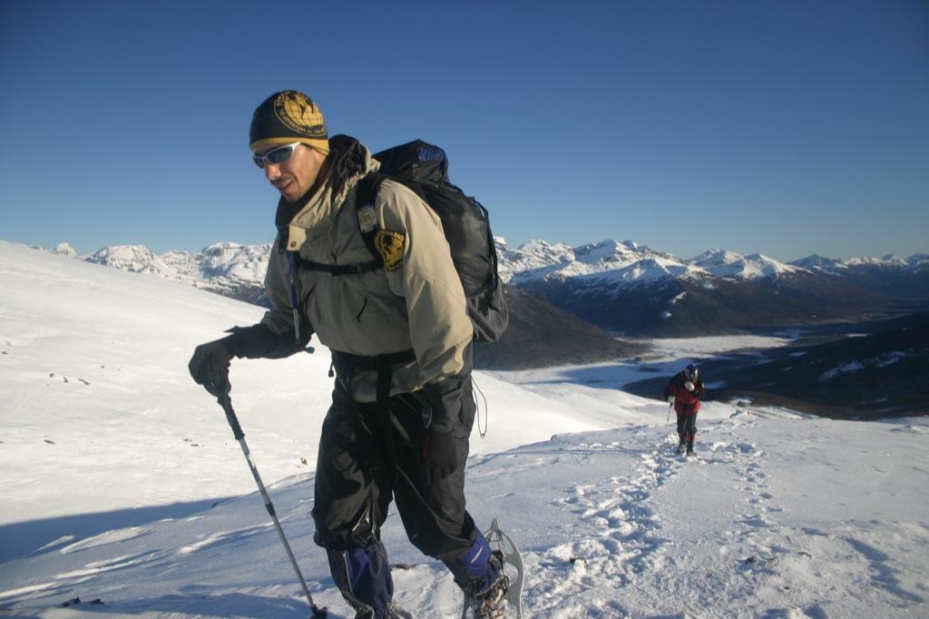 A side front landscape photo of two people snowshooing up a snowfield. In front is a man in full cold weather hiking gear. You can see him sweating in his face. On his head he is wearing a Original Buff® as beanie. The design is a custom design for the Patagonia Adventure Race. The other person is tracking about 50 metre behind. Source: buff.eu © Distributed for the promotion of the Original Buff® in outdoor / adventure