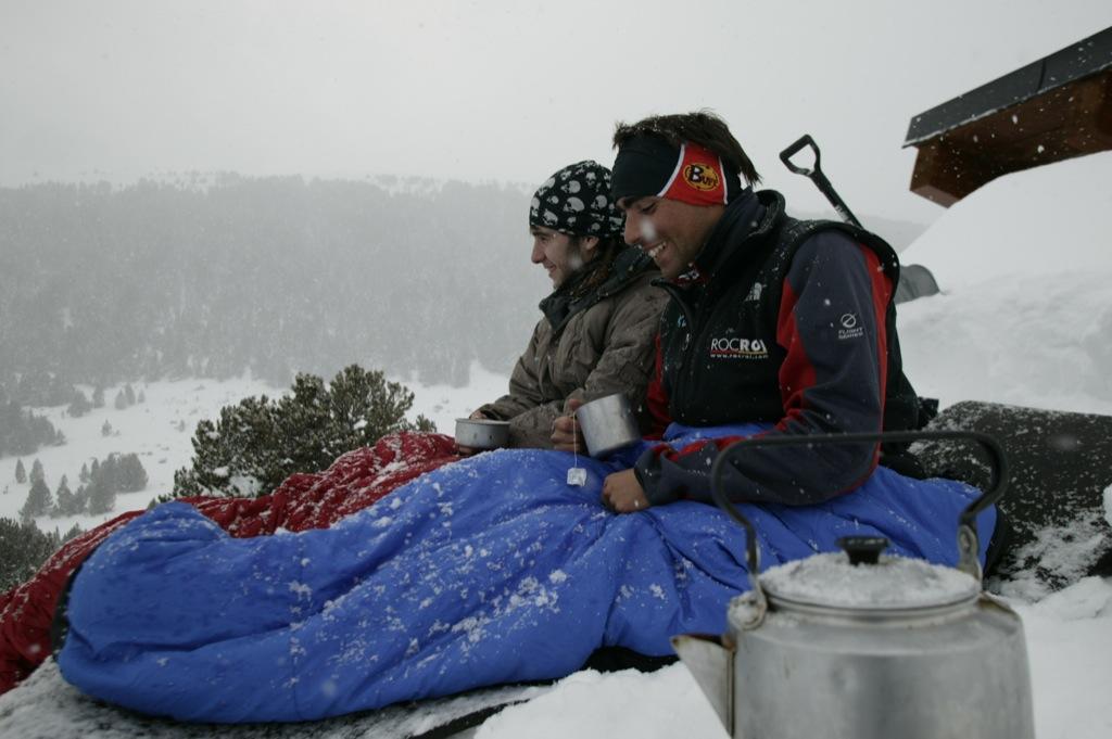 A side shot landscape photo of two men in the snow. They are sitting in their sleeping bags with their legs stretched out . It looks as if they sleeped outside and it started snowing. They are both holding a cup of tea in their hands and look happy. The man in front is wearing a Original Buff® as ear warmer. The man in the back is wearing a Original Buff® as beanie. Source: buff.eu © distributed for the promotion of the Original Buff® in outdoor / adventure