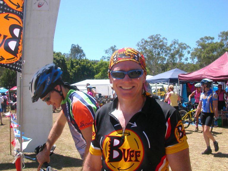 Landscape frontal shot photo of a male mountain bike rider. Scott smiling into the camera at the 2005 24 hour mtb race in Kooralbyne. It is hot. He is wearing a Original Buff® as helmet liner to cool the heat down. Source: Edward Copyright: ©2004 Buff Downunder Pty Ltd