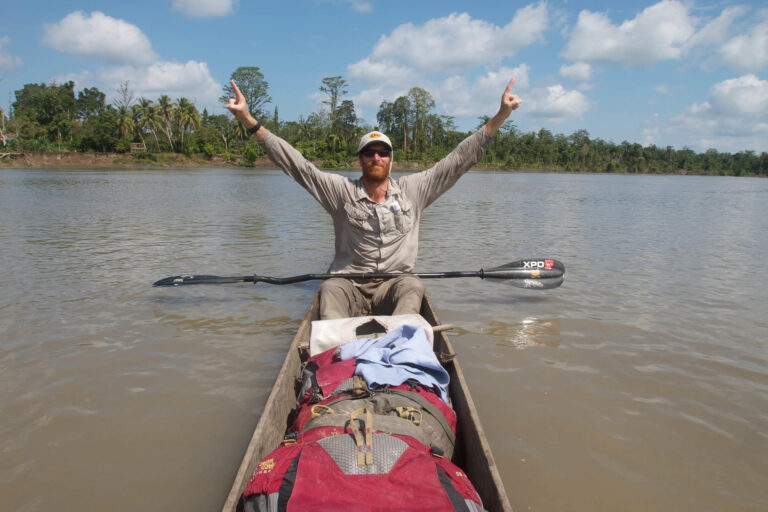 A frontal shot landscape photo of a man in a canoe raising his arms as a sign of victory. It is Andrew Johnson on the Sepik River in Papua New Guinea. He is wearing a Visor® Buff® as legionnaire style cap. The scene looks hot & humid. Image taken during the first spring to sea traverse of the Sepik River in Papua New Guinea. Source: Clark Carter © Clark Carter. https://www.adventureplaybook.com/ permission to use on our websites