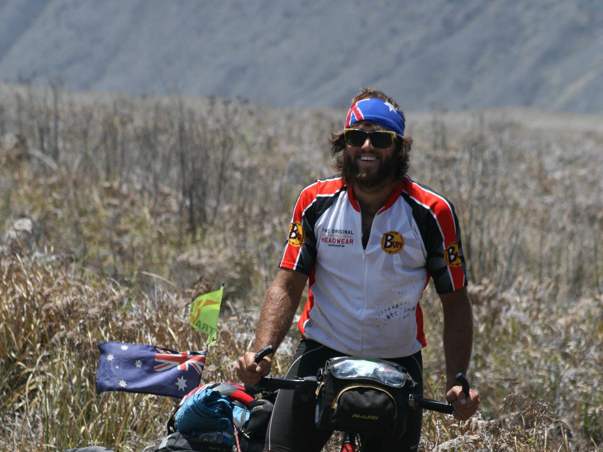 The horizontal image is a frontal shot of a man on a bicycle tour. It's Rian from EatSleepSurf.com. He has stopped in a field and is smiling at the camera. Rian is wearing Buff® team wear and a Australia Flag Original Buff®. It's obvious that non of the fabrics are wet or clingy. The location is in the highlands of Indonesia. It looks hot. But there are signs that there is also a chilly breeze. Rian is wearing his Original Buff® in layers. His flags on the bike are also flying. Source: EatSleepSurf.com.au Copyright: EatSleepSuf.com.au