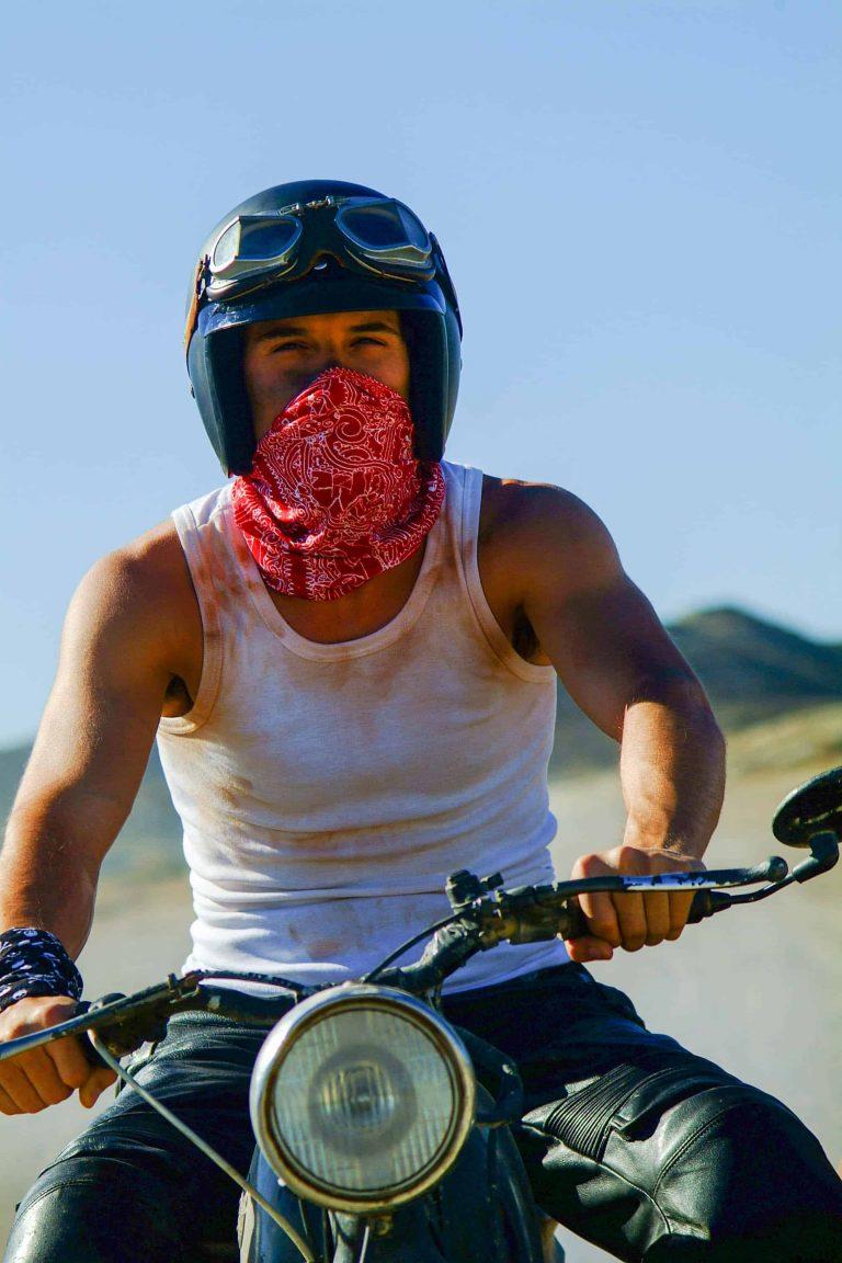 A frontal shot portrait photo of a man on a vintage motorbike. The man is wearing a leather bike trouser and a white singlet. Has a one High UV Buff® as wristband on and one High UV Buff® as face mask. He is also wearing an open face motorbike helmet and vintage motorcycle goggles over the helmet. The background looks hot & dusty. Source: buff.eu Copyright: Distributed for the promotion of the High UV Buff® in the motorcycling