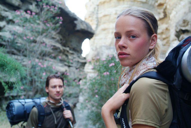 A side landscape shot of a woman and a man hiking in a gorge. Both are wearing Original Buff® as neck cooler. The woman to the front right is looking over her left shoulder into the camera. The man in the background seems distracted. The scene looks warm. Both are wearing summer hiking gear. Source: buff.eu © distributed for the promotion of the Original Buff® in hiking / trekking