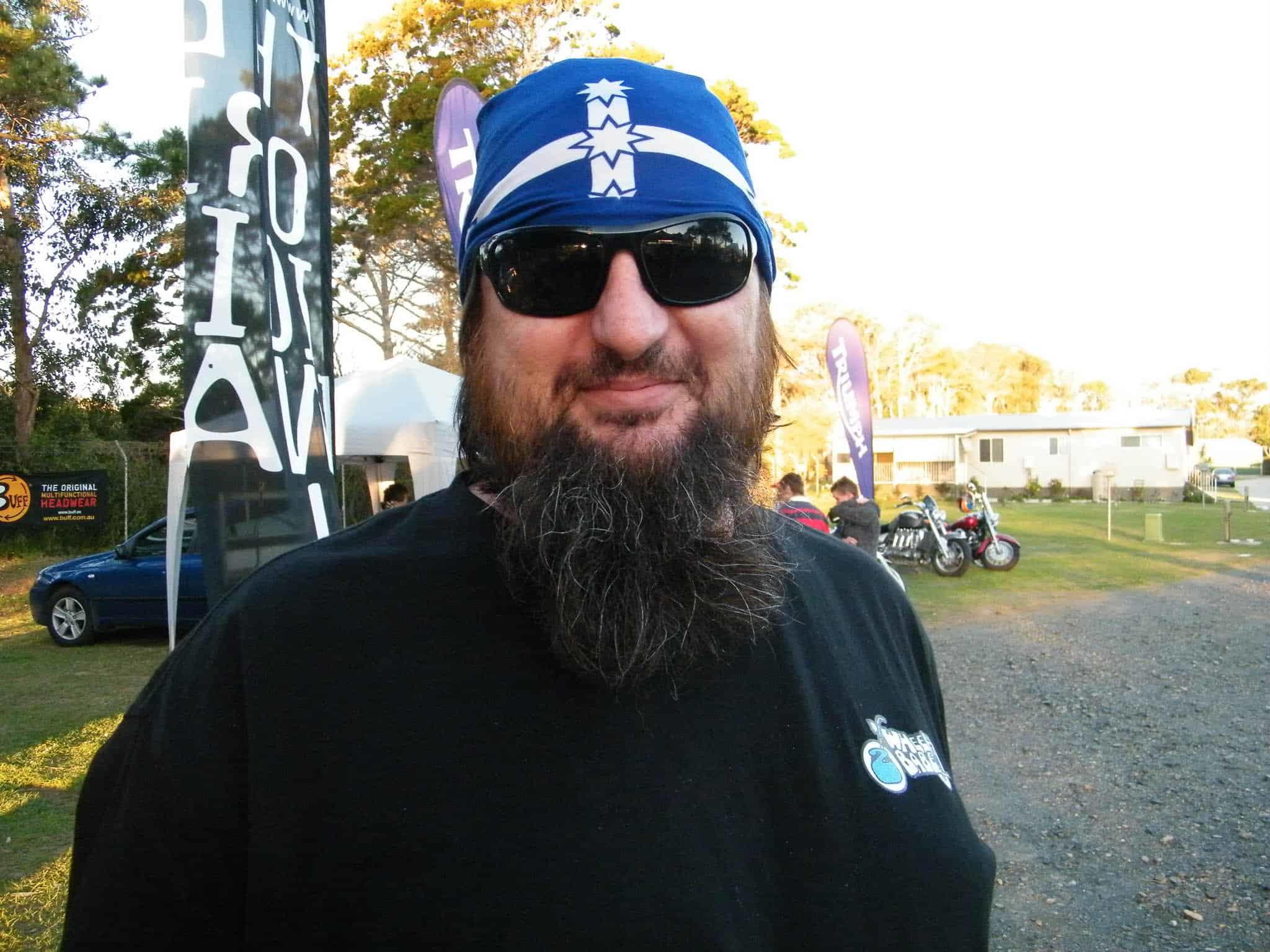 A landscape upper body shot of a bearded man looking straight into the camera. He sports a long beard, dark sunglasses & a Eureka Flag Original Buff®. He wears the Buff® as a bandana / pirate cap with the Eureka flag prominently on his forehead. He clearly identifies as a biker on a motorbike event. The photo was taken on the 2010 Wheel Babes Meeting in Ballina, NSW, Australia. The man was helping out as volunteer. Source: Edward Copyright: ©2010 Buff Downunder Pty Ltd
