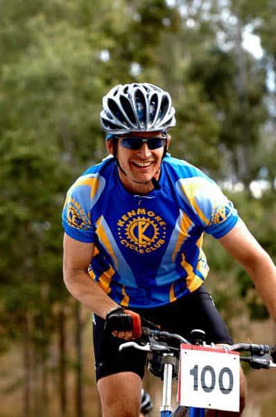 The image shows John from Kenmore Cycles during the 2004 Kooralbyne 24 hour MTB XC race. He is wearing a Original Buff® as legionnaire cap under his helmet. This keeps the sweat out of his eyes and gives heat relief. I mention the High UV Buff® in the caption because today this would be your preferred product for this situation. The High UV Buff® excels in handling sweat & heat. The photo is a vertical shot showing John from the handlebars upwards. John is smiling and it looks as if he is having a great time. He is wearing mountain bike gloves, a short sleeved "Kenmore Cycle Club" cycling jersey, blue tinted sunglasses and a helmet with big vents. The Buff® is clearly visible under the helmet. Photographer: Unknown Copyright: Unknown but we received the images as part of being sponsors with the right to use commercially