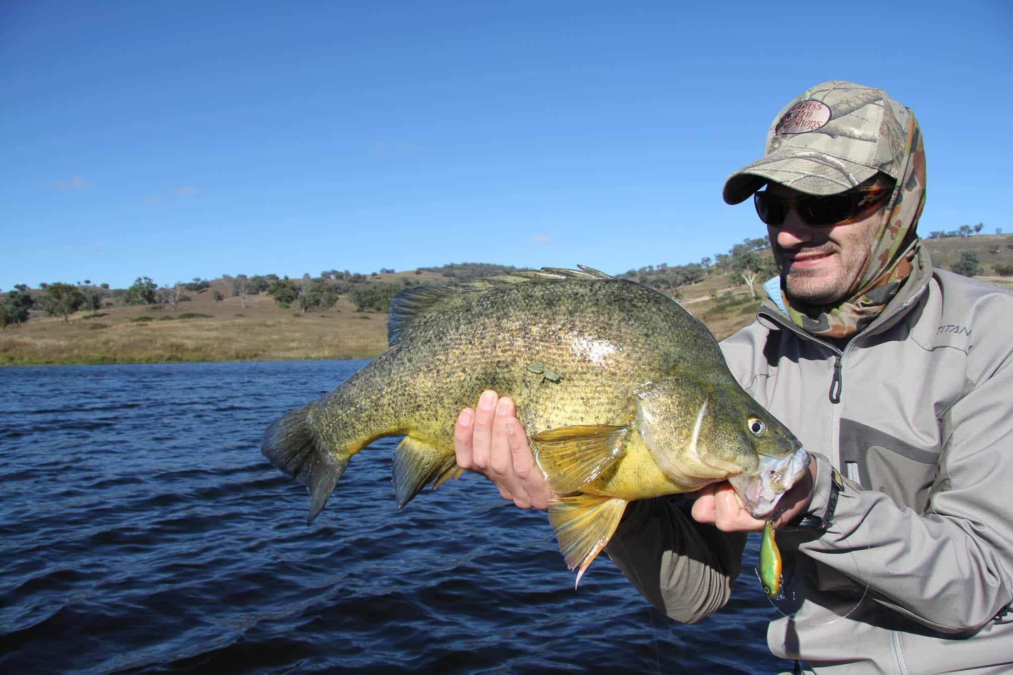 A landscape front shot of a man holding up a large fish. The fish is in the centre of the frame and the man is standing to the right. He is admiring his catch and smiling. The weather looks sunny but cold. The man is wearing a cold weather jacket and a Auscam Original Buff® as scarf / half balaclava. Source: Testimonial Copyright: Permission to use on our website