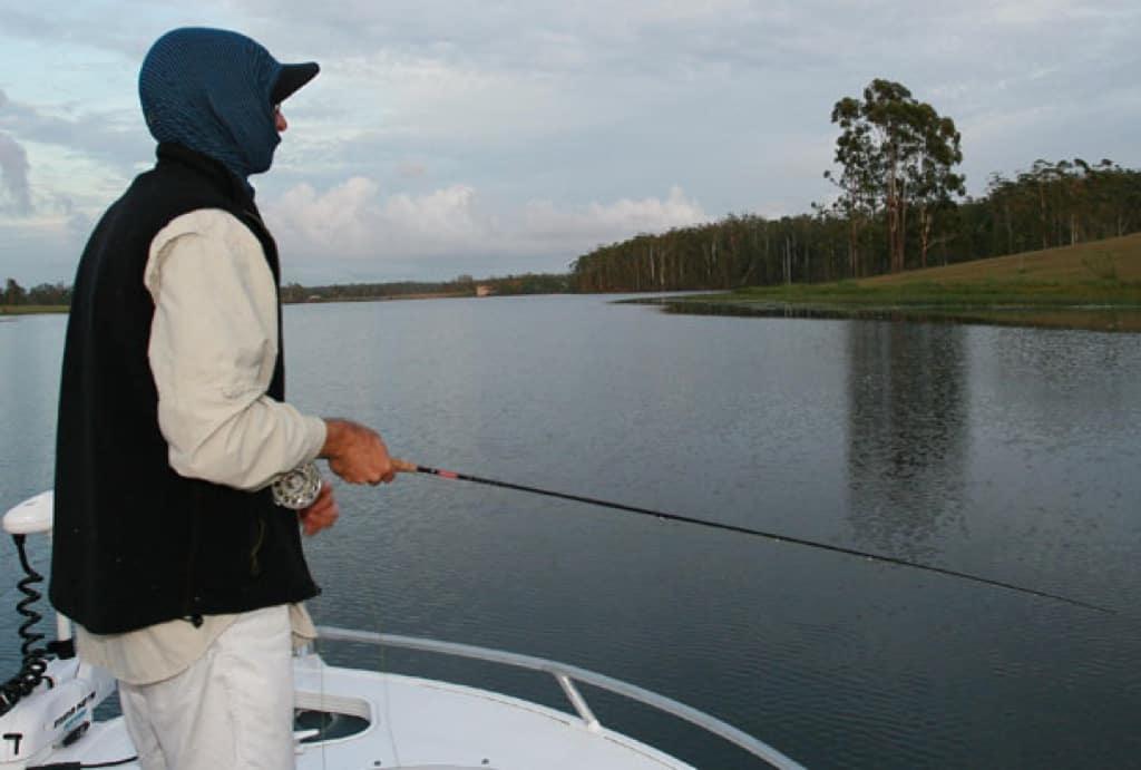 A landscape photo of a person fly fishing on a lake. The person is standing on a boat wearing a Visor Buff® as full Balaclava. The day looks cool to cold. Full cloud cover and the person is wearing a fleece west. Source: Testimonial Copyright: Permission to use on our website