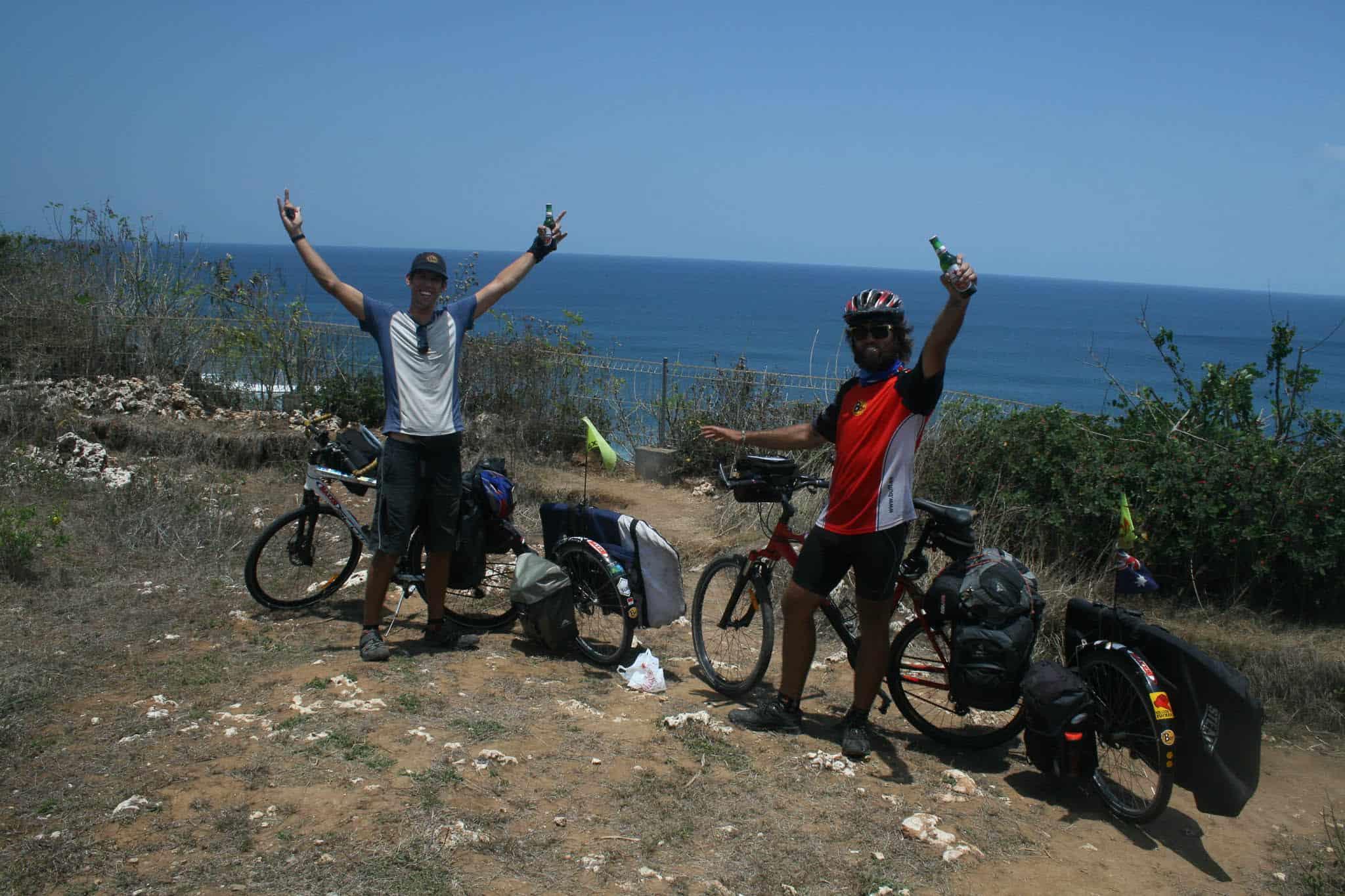 The horizontal photo shows Rian and Dylan standing with their bicycles on the cliff of Uluwatu, Bali, Indonesia. They have cycled Indonesia from North to South and are celebrating their successful journey. Dylan is holding both arms up as a victory sign. Rian has got one arm up. He is holding a beer. Rian is wearing the Australia Flag Original Buff® as neck cooler. It appears mid day with hardly any shadow and a clear blue sky. It must be hot. Source: Eatsleepsurf.com.au Copyright: Unknown. We received the images as "Thank You" for providing the Buff®