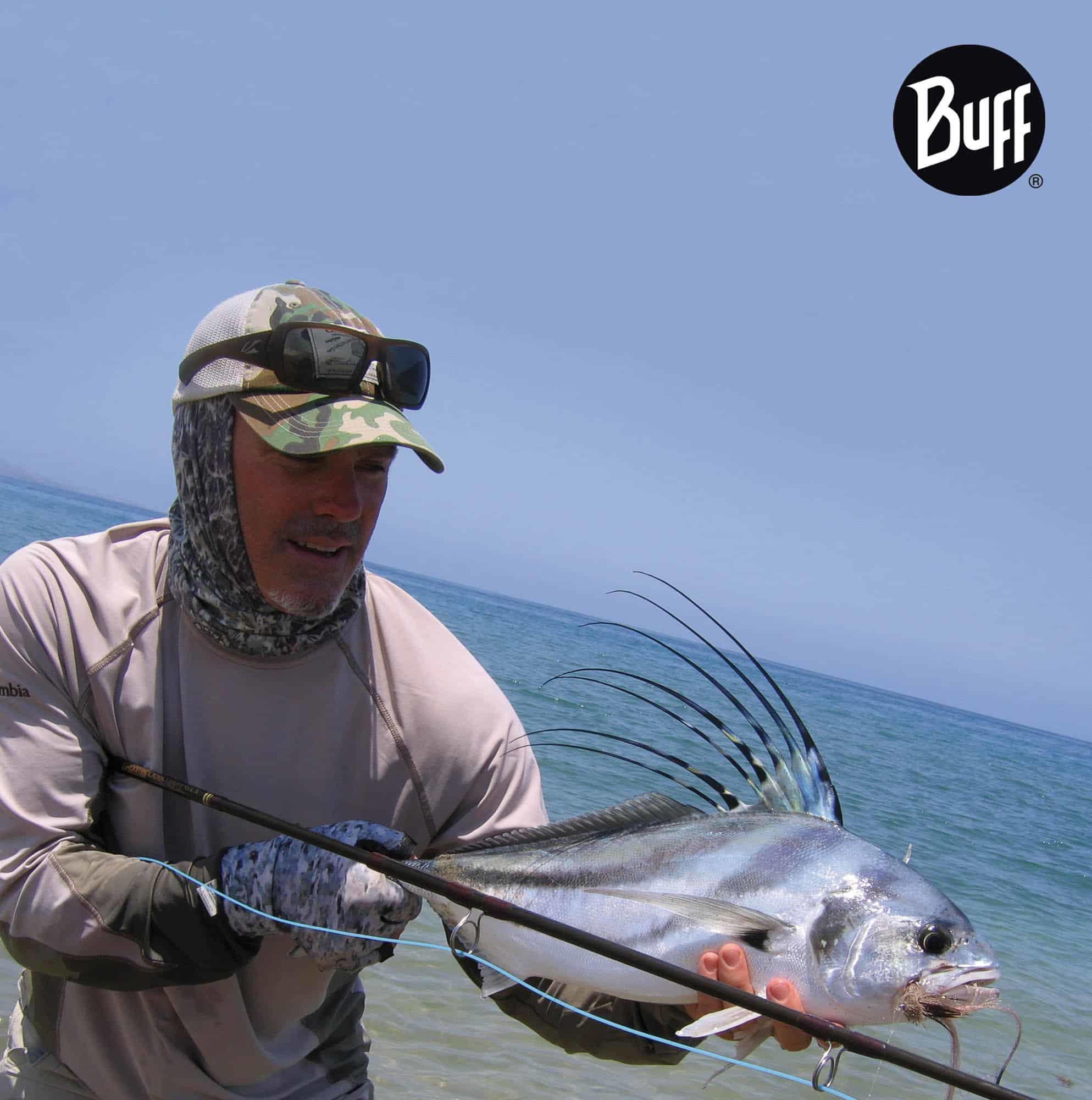 The photo shows a saltwater fisherman holding up a great catch. He is smiling at his catch. He is wearing a High UV Buff® and Angler Gloves. The scene looks hot & sunny. The man is wearing the High UV Buff® as a half balaclava. The face is entirely exposed. This and the shadows indicates that the sun is behind the man. Source: buff.eu Copyright: Unknown. Distributed for the promotion of the HIgh UV Buff® in fishing.