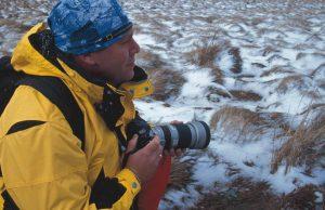 A side shot landscape photo of a man knealing in the snow. It looks like he is on a photo safari or researching wildlife. He is wearing a yellow cold weather ourtdoor jacket. He is also wearing a Original Buff® as beanie. The design is clearly recognisable as a National Geographing Collection design. Source: buff.eu © Distributed for the promotion of the Original Buff® in outdoor / adventure