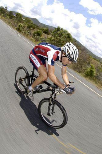 A vertical photo of a male riding a XC mountain bike in the centre of a bitumen road. The rider is wearing a red & blue Simplon team jersey and cycle shorts. Under his white mtb helmet you can spot the Crocodile Trophy High UV Buff®. He is wearing it as a legionnaire cap covering his head and his back neck. The speed is fast paced and the surroundings looks like the can fields north of Port Douglas in North Queensland. Source: Crocodile Trohpy Photographer unknown Copyright: Unknown. We received this low-res images as "Thank-You" for providing the High UV Buff®