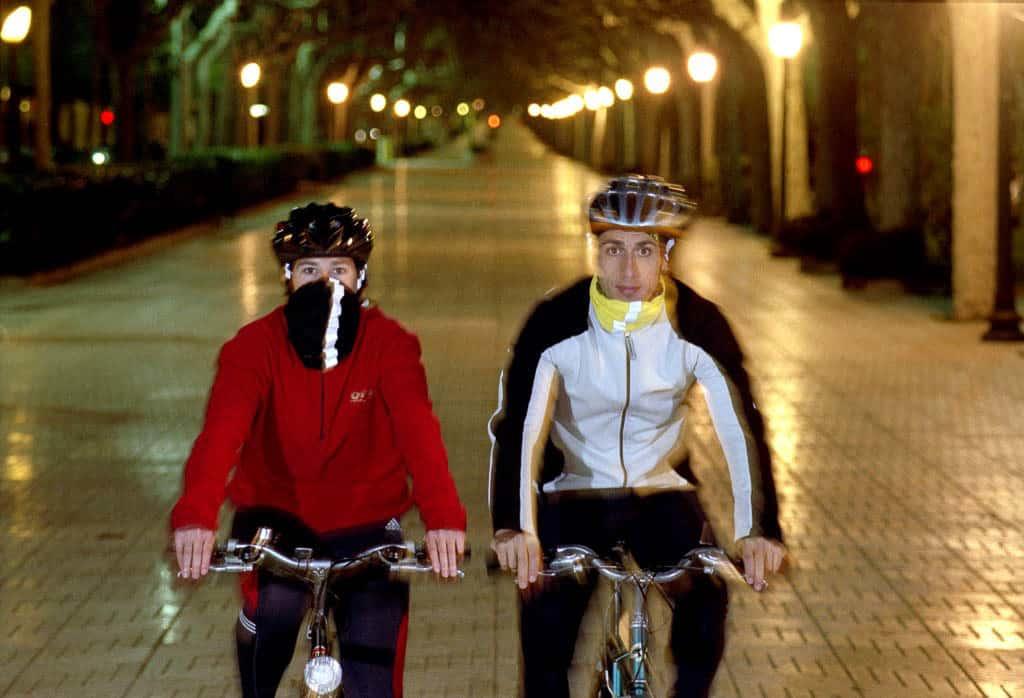 The horizontal photo is a frontal shot of a woman and a man riding bicycles in a city mall. It is dark and both are wearing cold weather gear. The woman on the left is wearing a Reflective Buff® as face mask. The man on the right is wearing a Reflective Buff® as scarf. Source: buff.eu Copyright: Unknown. Distributed for the promotion of the Reflective Buff®