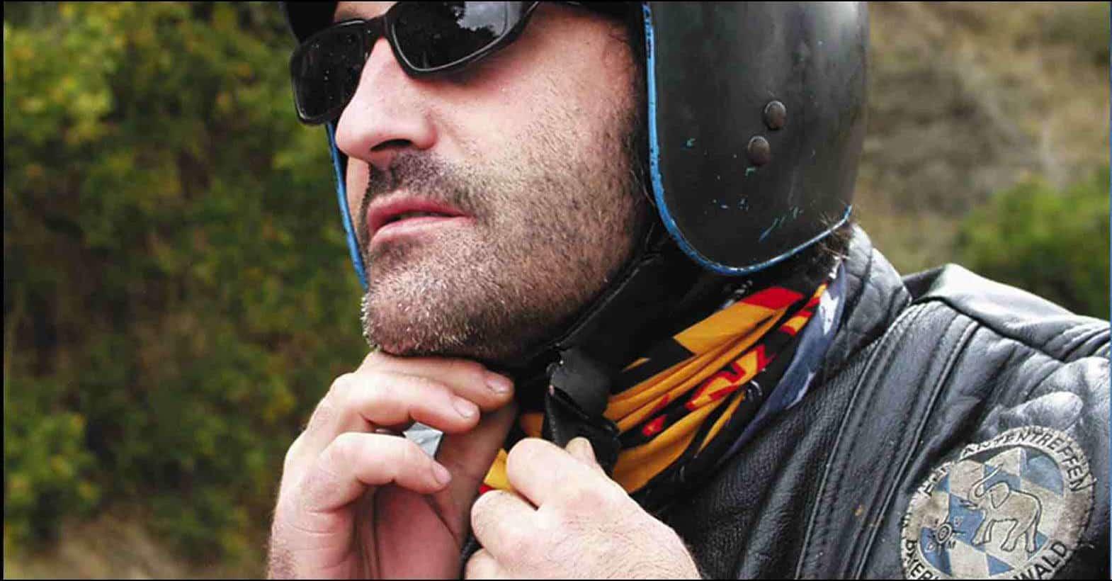 A side landscape shot of a man tying his motorcycle helmet. Visible is his upper body with the top of his helmet not visible. He is wearing a motorcycle leather jacket and a Original Buff® as scarf. His helmet is matt black and he is also wearing black sunglasses. He looks like a Harley rider. Interesting in this photo is how nice the Buff® closes up the jacket. It fills out all the nooks and crannies. The weather looks classic European summer. Not hot & not cold with a slight chill in the morning. Source: buff.eu Copyright: Distributed for the promotion of the Original Buff® in motorcycle