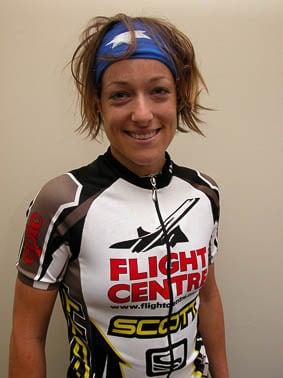 The photo is vertical and shows female MTB rider Tory standing upright in front of a grey wall. She is wearing a Flightcentre Scott team cycle jersey and a Original Australia Flag Buff® as Alice Band. Her tired smile and her hair suggest that the photo is taken after a XC race. She smiles tired but happy. The Buff® as Alice Band keeps her hair together and tidy. Photographer: Unknown Copyright: Unknown. We received the image as a "Thank-You" for providing the Flight Centre Scott MTB team with Australia Flag Buff®