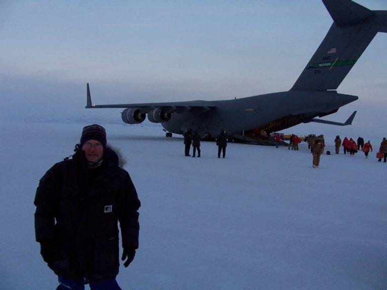A front landscape shot of a man standing on an ice field in front of a large military supply air craft. The back loading dock is open and people are moving around the plane. It is the runway of the Scott Base in Antarctica. The man is wearing a one piece down overall with a fur hood. Around his neck is a Original Buff® as scarf. Source: Michael Nottage © permission to use on our website