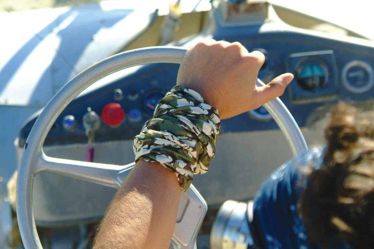 Man driving a car. Photo is taken from behind. It shows the left arm holding the steering wheel. A High Uv Buff® as wristband is the main element. Photoshoot for Buff® summer catalogue with models. © Original Buff S.A
