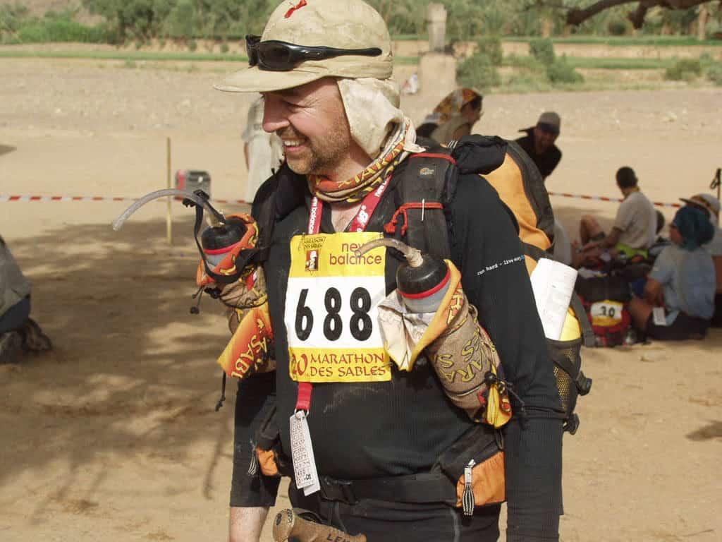 A side, landscape shot of a man smiling in full Marathon Des Sables gear. He is wearing a Original Buff® as neck cooler. He looks happy & relaxed. I assume that this photo was taken before a stage of the 2005 Marathon Des Sables. You can also spot other uses of the Original Buff® like water bottle covers. Today people prefer the High UV Buff® for this event. Source: buff.eu © distributed for the promotion of the Original Buff®.