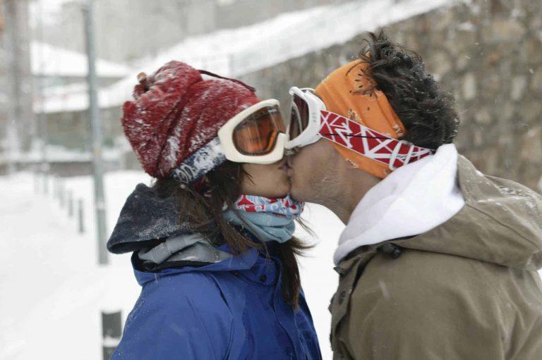 A landscape shot of a couple kissing. It is obvious that they are on ski holidays. She is wearing a Polar Buff® as scarf and a Neckwarmer Buff® as hat. He is wearing a Original Buff® as ear warmer. Source: buff.eu. Distributed for the promotion of Buff® products