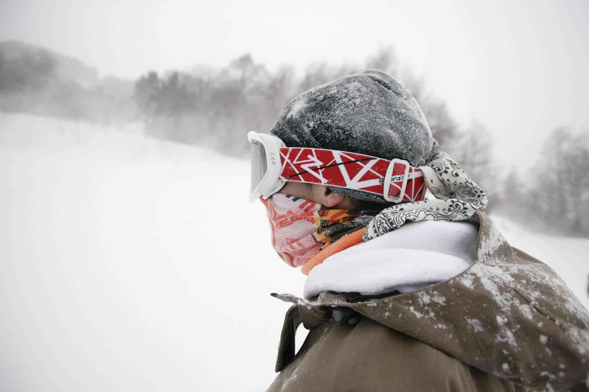 A landscape shot of a freestyle snowboarder. The conditions are rough. Strong winds are blowing snow around. He is wearing two Polar Buff®. One as scarf & face mask combination. The other one as hat / beanie. Source: buff.eu. Distributed for the promotion of the Polar Buff®