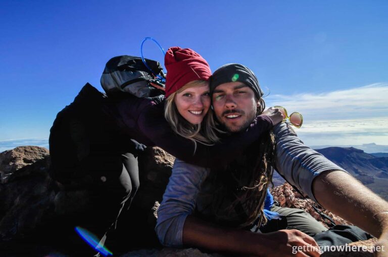A landscape selfie showing a young man and woman smiling in the camera. They are sitting on top of a mountain peak. Their faces reveal exhaustion and happiness. They are both wearing Wool Buff® as beanies. Source: Gettingnowhere.net © Permission to use on our websites