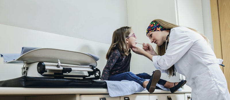 Photo of a female doctor examining a young girl. The doctor is wearing a Medical Collection Dry Cool Buff® in bright colours. The photo could be taken in a hospital or a medical practise. It looks white & sterile. Source: buff.eu