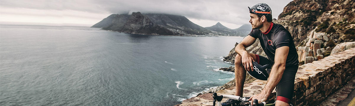 A wide format image of a male cyclist stopping at the side of the road and looking at the scenery. You can see a large body of water and an island in the background. The man is leaning against a rock wall type road boundary. Source: buffusa.com