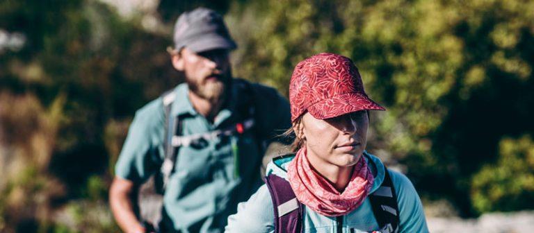 A catalogue hero photo of a man and a woman hiking. They are wearing cool environment clothing with a Buff® around the neck and a Pack Treck Cap on the head. The woman in the foreground is sharp and in focus. The man in the back and the background are blurry. Source: buff.eu