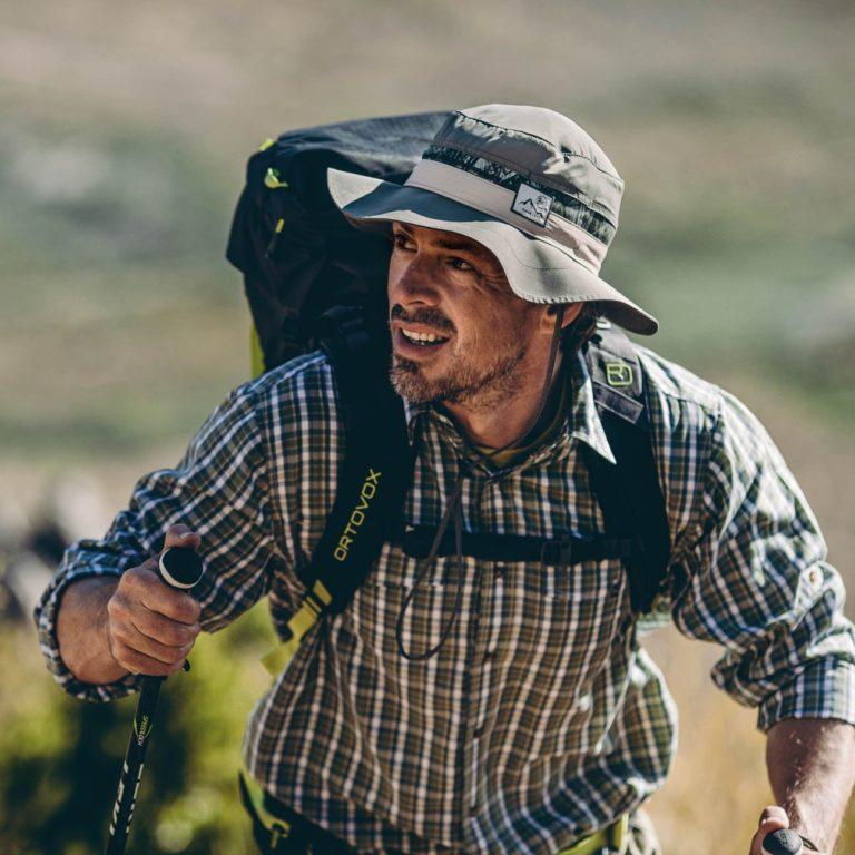 Picture of a man hiking up a hill. It looks sunny and hot. The man is wearing a Buff® Booney Hat and is carrying a backpack. He is also using walking poles. Source: buff.eu