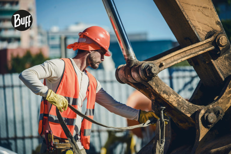 The image shows a construction worker using a Dry Cool or a Modacryl FR Buff® as helmet liner. The man is working on lifting gear with heavy machinery in the background. A Buff® trademark logo is placed on the top left corner. Source: buff.eu