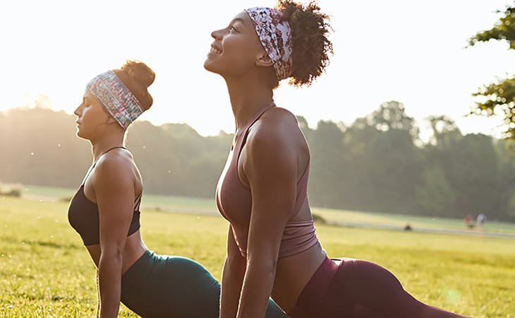 2 women performing a yoga pose in the park. They are both wearing the BUFF® Coolnet UV+ Tapered Headband as sweatband to tame their long and curly hair. Source: buff.eu