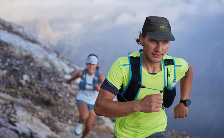 A man and a woman running up a mountain. It looks as if they are ultr-running for fun as they both look very happy. The man in the foreground is wearing a BUFF® Pack Speed Cap. The woman in the background is wearing a BUFF® visor. Source: buff.eu
