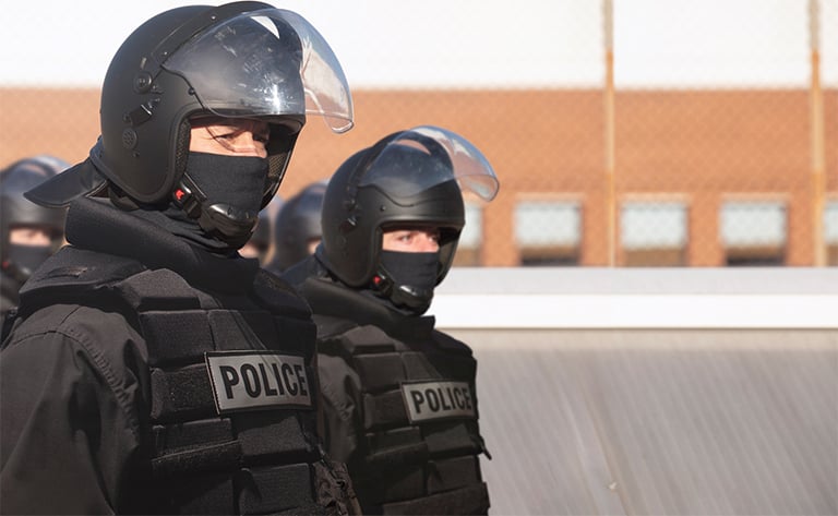 A photo of a male and female police officer in full urban warfare gear. They are both wearing a black BUFF® Safety Fire Resistant Balaclava under their helmets. The Balaclava covers their face all the way up to their eyes. Source: buff.eu